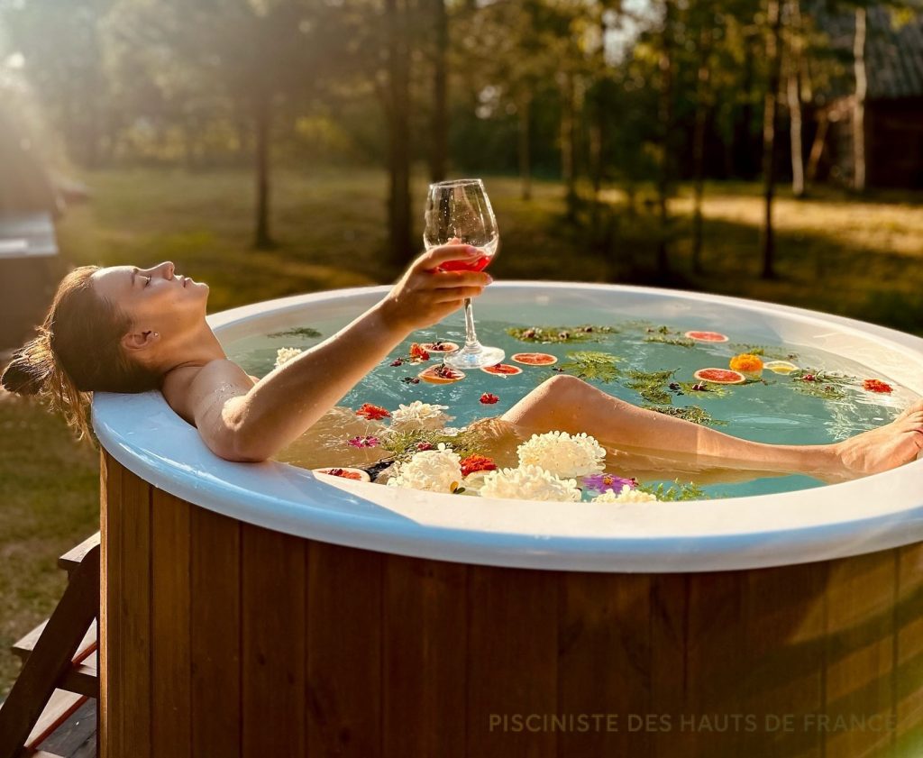 Femme se relaxant dans un bain nordique PolarSpa avec un verre de vin et des fleurs flottantes dans un jardin des Hauts-de-France.