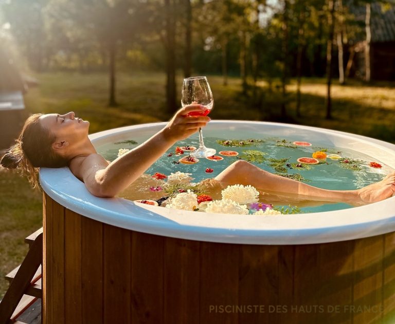 Femme se relaxant dans un bain nordique PolarSpa avec un verre de vin et des fleurs flottantes dans un jardin des Hauts-de-France.